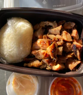 Grilled Thai chicken pieces with a ball of sticky rice and two dipping sauces in a black to-go tray