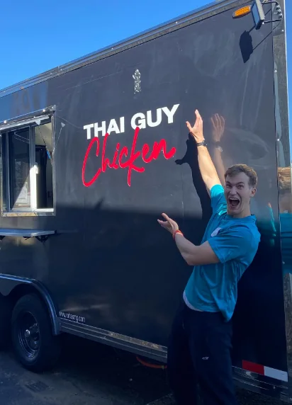 Jake Pennington grinning and pointing at the Thai Guy Chicken logo on the side of the black food truck